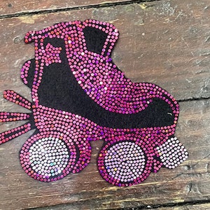 May include: A decorative roller skate applique. The skate is black with pink rhinestones. The wheels and other details are also pink with rhinestones. The applique is on a wooden surface.