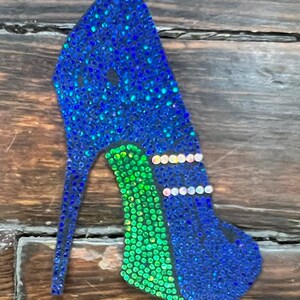 May include: A high-heeled shoe-shaped ornament covered in sparkling blue and green rhinestones. The heel is a thin, pointed design. The shoe features a green arch and is decorated with rows of white rhinestones.