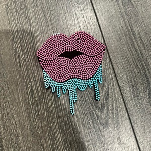 May include: A pink and blue rhinestone lips design. The upper lip is pink, and the lower lip is dripping with blue rhinestones. The lips are made of small, round rhinestones.