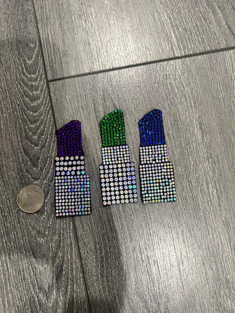 Pu&ograve; includere: Tre accessori a forma di rossetto ricoperti di strass, in viola, verde e blu. Sono disposti su una superficie di legno grigio. Una moneta &egrave; nell'immagine per il confronto delle dimensioni.