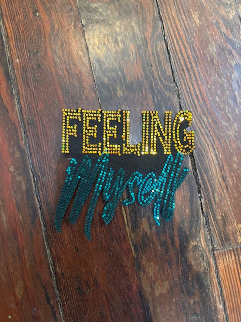 May include: A decorative piece with the words "FEELING Myself" spelled out in gold and teal sequins. The gold letters are in a block style, while the teal letters are in a cursive style. The piece is set against a dark wood background.