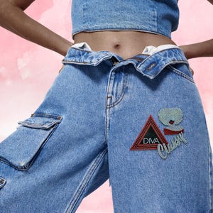 Pode incluir: Calças cargo de ganga azul com cós dobrado e um emblema decorado com strass na coxa esquerda. O emblema inclui um triângulo com rebordo vermelho com o texto DIVA, uma figura estilizada e a palavra Classy em escrita prateada.