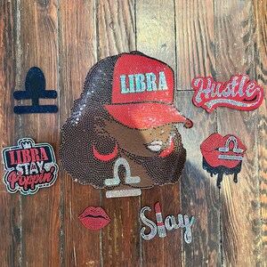 Könnte beinhalten: Eine Sammlung von Accessoires zum Thema Waage mit Strasssteinen. Enthält ein Porträt einer Frau mit roter Kappe mit "LIBRA", zusammen mit den Wörtern "Hustle" und "Stay" und anderen Waage-Symbolen. Auf Holzgrund.