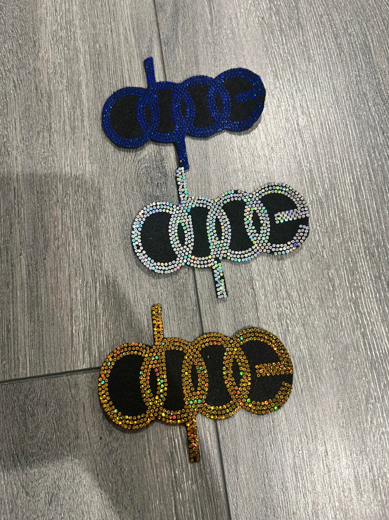 Peut inclure: Trois d&eacute;corations de g&acirc;teau avec le mot "ONE". Les d&eacute;corations sont bleues, iris&eacute;es et dor&eacute;es. Chaque "ONE" est bord&eacute; de strass et a un fond noir. Les d&eacute;corations sont pos&eacute;es sur une surface en bois grise.