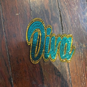 May include: A bedazzled Diva brooch. The word "Diva" is spelled out in teal and gold rhinestones. The brooch is on a wooden surface.