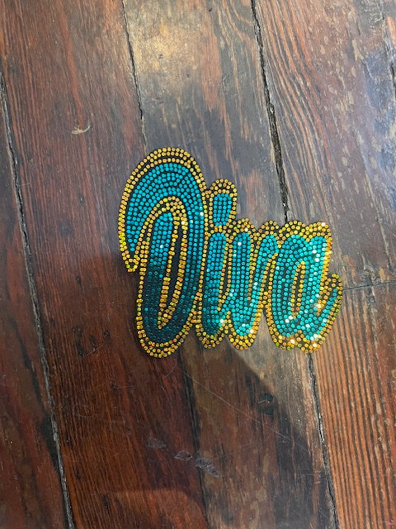 May include: A bedazzled Diva brooch. The word "Diva" is spelled out in teal and gold rhinestones. The brooch is on a wooden surface.