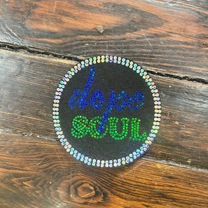 May include: A round, black patch with the words "DOPE SOUL" in blue and green rhinestones. The patch is encircled by a border of iridescent rhinestones. The patch is set against a wooden background.