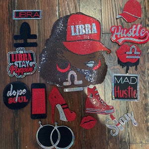 May include: A collection of rhinestone-studded patches and appliques, including a portrait wearing a red "LIBRA" cap, a red high-heeled shoe, a red sneaker, and the word "Hustle". The items are arranged on a wooden surface.