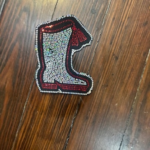 Puede incluir: Un parche en forma de bota adornado con pedrería. La bota tiene un contorno negro, una parte superior roja y detalles. El cuerpo de la bota está relleno de pedrería iridiscente, y los bordes están bordeados con pedrería blanca.