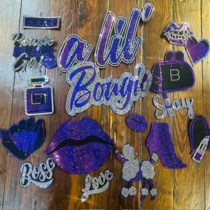May include: A collection of rhinestone-encrusted patches and appliques in purple, silver, and black. Designs feature text such as "Bougie," "Boss," and "Slay," alongside images of lips, a perfume bottle, a purse, and a poodle. The patches are displayed on a wooden surface.