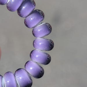May include: A circular hair coil accessory featuring a smooth, translucent purple gradient finish. The material transitions from a lighter lavender hue to a deeper, saturated violet tone, creating a glossy, reflective surface across each individual loop.