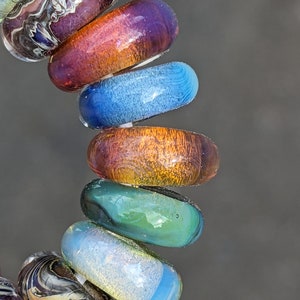 Puede incluir: Una pila de cuentas de vidrio de colores con patrones en espiral. Las cuentas están dispuestas en orden de arcoíris, comenzando por el morado y terminando por el azul.