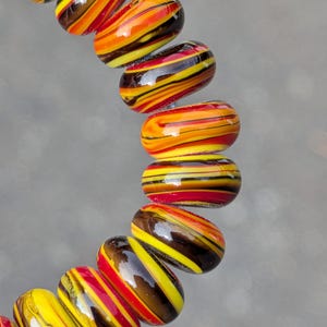 May include: A circular arrangement of smooth, cylindrical glass beads featuring a marbled pattern of vibrant yellow, bright orange, deep red, and dark brown horizontal stripes. The beads have a glossy, reflective finish and are strung together.