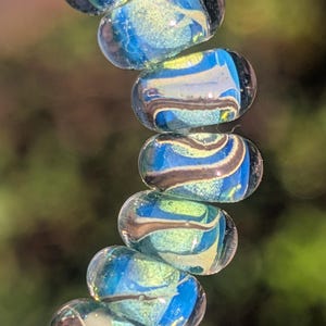 Puede incluir: Primer plano de un collar de cuentas de vidrio. Las cuentas son de forma ovalada con una base transparente y presentan remolinos de azul, verde y marrón. Las cuentas están ensartadas, creando un accesorio decorativo.