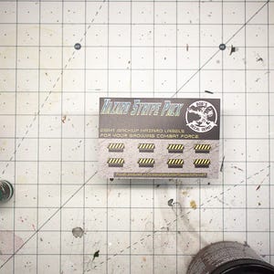 May include: A top-down shot of a crafting workspace with a grid-patterned surface. The focus is on a package labeled "Hazard Stripe Pack" with yellow and black hazard stripe decals. Other items include paint, a brush, and wooden components.
