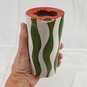 Könnte beinhalten: Eine Keramikvase mit Wassermelonen-Design. Der obere Rand ist rot, der Körper ist weiß mit grünen Wellenstreifen. Die Vase wird in einer Hand vor weißem Hintergrund gehalten. Die Vase ist etwa 15 cm hoch.