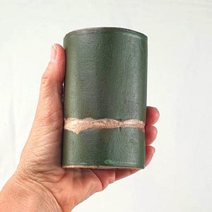 Könnte beinhalten: Eine zylindrische, handgefertigte Keramikvase in einem tiefgrünen Farbton. Die Vase hat ein helleres, strukturiertes Band in der Mitte. Das Gesamtdesign ist schlicht und rustikal, passend für die Inneneinrichtung.