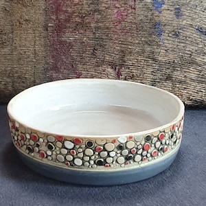 Könnte beinhalten: Eine kleine, weiße Keramikschüssel mit einem dekorativen Rand. Der Rand ist mit einem Muster aus kleinen, bunten Kreisen in Rot-, Schwarz-, Weiß- und Beigetönen verziert. Die Schüssel hat einen grauen Boden.