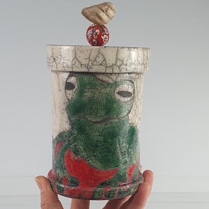 Könnte beinhalten: Ein Keramikbehälter mit einem grünen Froschmotiv. Der zylindrische Behälter hat eine Craquelé-Oberfläche und rote Akzente. Der Deckel ist mit einem roten Ball und einem kleinen Stein gekrönt. Ein einzigartiges dekoratives Objekt.
