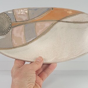 Könnte beinhalten: Eine ovale Servierplatte aus Keramik mit weißem Überzug und einem dekorativen Design in Grautönen, Rosa und Orange. Das Design zeigt eine stilisierte Blume mit einer spiralförmigen Mitte.