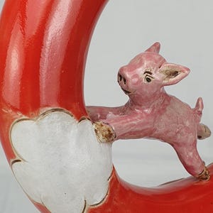 Könnte beinhalten: Eine Keramikskulptur, die ein rosa Ferkel zeigt, das sich an einem roten, ringförmigen Objekt mit weißen, wolkenartigen Akzenten festhält. Das Ferkel ist in Bewegung dargestellt, was dem Kunstwerk eine dynamische Qualität verleiht.