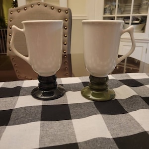 Op de afbeelding: Twee witte keramische mokken met unieke handvatten en decoratieve voetstukken. Eén mok heeft een zwarte voet, de andere een groene. De mokken staan op een zwart-wit geblokte tafelkleed.