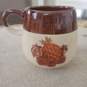 May include: A brown and white ceramic mug with a pineapple and strawberries design. The mug has a brown rim and handle.