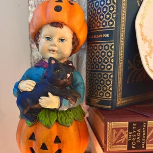 May include: A ceramic Halloween figurine of a child wearing a pumpkin hat and holding a black cat. The child stands on a carved pumpkin with a smiling face. The figurine is surrounded by books with gold lettering.