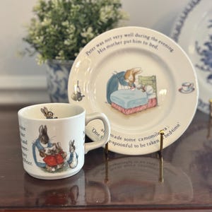 May include: A white ceramic mug and plate set featuring illustrations of Peter Rabbit. The mug has a handle and text, while the plate depicts Peter in bed. The set is displayed on a wooden surface.