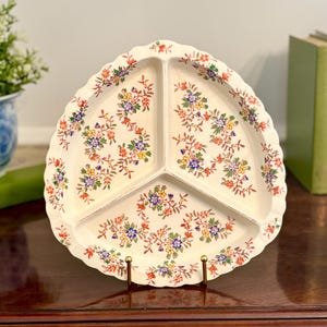May include: A cream-colored ceramic divided serving dish with a scalloped edge and a three-section interior. The surface is decorated with a repeating floral pattern featuring small blue, yellow, and orange flowers with green leaves and brown stems.