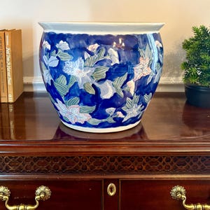 May include: A large, round ceramic planter featuring a deep blue glazed background decorated with a repeating pattern of light pink and white flowers, green leaves, and stems. The bowl-shaped vessel has a wide, flared rim and a smooth finish.