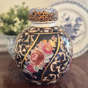 May include: A black and gold ceramic jar with a lid, featuring floral and scrollwork designs. The jar has a round shape and is adorned with pink, red, and yellow flowers. The lid has a blue and brown pattern. A decorative piece for the home.