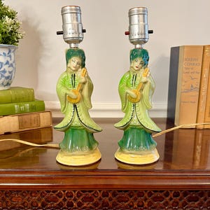 May include: A pair of ceramic table lamps featuring figures in green robes playing stringed instruments. Each lamp has a light green and dark green glazed finish, a yellow base, and a silver metal socket at the top for a light bulb.