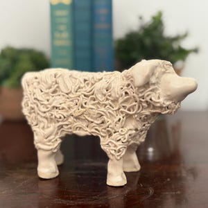 May include: A cream-colored ceramic sheep figurine with a textured, curly wool design. The sheep has four legs and a detailed face. The figurine is a decorative item, likely for home decor.