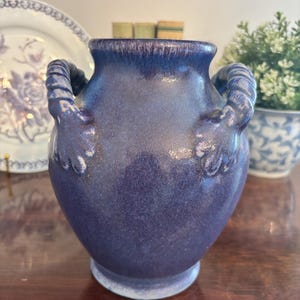 May include: A dark blue ceramic vase with two rope-like handles. The vase has a rounded body and a flared rim. The glaze has a textured appearance. Decorative item for home decor.