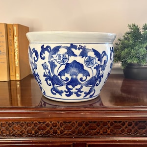 May include: A white ceramic planter featuring a repeating blue floral and scrollwork pattern. The bowl-shaped vessel has a wide, smooth rim and a tapered base, decorated with intricate botanical motifs in a deep cobalt blue glaze.