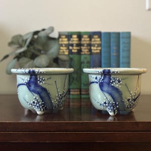 May include: Two matching ceramic pots with a light green base and blue painted design. The pots have a rounded shape with three small feet and a decorative rim. White dots accent the blue design.