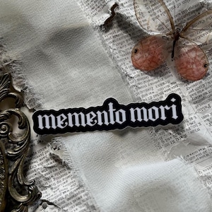 Peut inclure: Autocollant noir et blanc avec le texte "memento mori" en police gothique. L'autocollant est sur un fond blanc avec un morceau de papier d&eacute;chir&eacute; et un papillon.