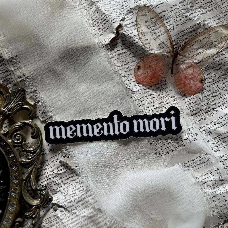 Peut inclure: Autocollant noir et blanc avec le texte "memento mori" en police gothique. L'autocollant est sur un fond blanc avec un morceau de papier d&eacute;chir&eacute; et un papillon.