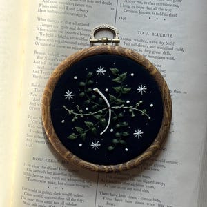 Peut inclure: Un cercle brodé avec un fond bleu foncé et un cadre en bois. La broderie présente un arc blanc et des feuilles vertes avec des étoiles blanches. L'œuvre est présentée sur un livre ouvert.