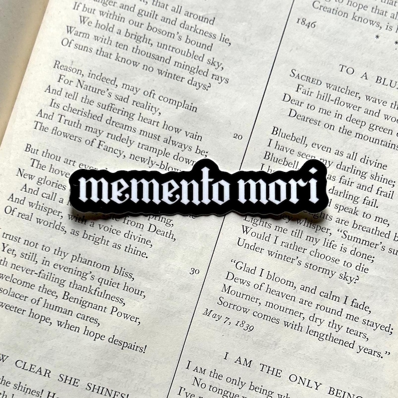 Peut inclure: Autocollant noir et blanc avec le texte "memento mori" en police gothique. L'autocollant est sur une page d'un livre avec du texte sur la nature et la mort.