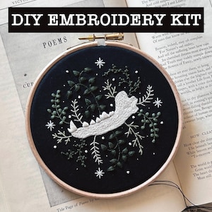May include: A DIY embroidery kit featuring a circular wooden hoop containing black fabric. The embroidery depicts a white animal jawbone surrounded by dark green botanical vines, leaves, and small white star-shaped accents.
