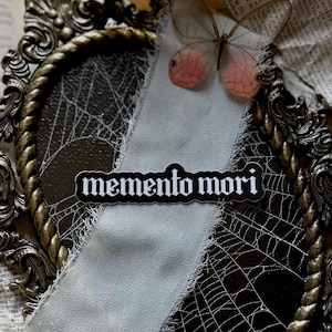 Peut inclure: Un autocollant noir et blanc avec le texte "memento mori" dans une police gothique. L'autocollant est plac&eacute; sur un tissu blanc aux bords effiloch&eacute;s, qui est drap&eacute; sur un cadre orn&eacute; dor&eacute; avec un motif de toile d'araign&eacute;e. Un d&eacute;licat papillon aux ailes roses est visible en arri&egrave;re-plan.