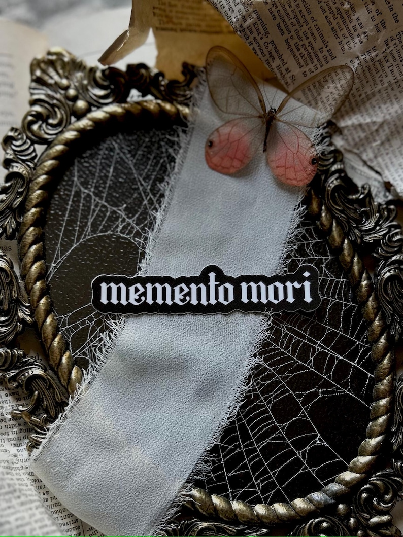 Peut inclure: Un autocollant noir et blanc avec le texte "memento mori" dans une police gothique. L'autocollant est plac&eacute; sur un tissu blanc aux bords effiloch&eacute;s, qui est drap&eacute; sur un cadre orn&eacute; dor&eacute; avec un motif de toile d'araign&eacute;e. Un d&eacute;licat papillon aux ailes roses est visible en arri&egrave;re-plan.