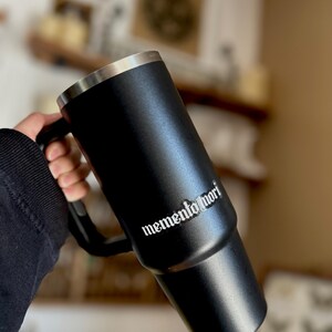 Peut inclure: Mug de voyage en acier inoxydable noir avec une poign&eacute;e noire et une inscription blanche qui dit "memento mori".