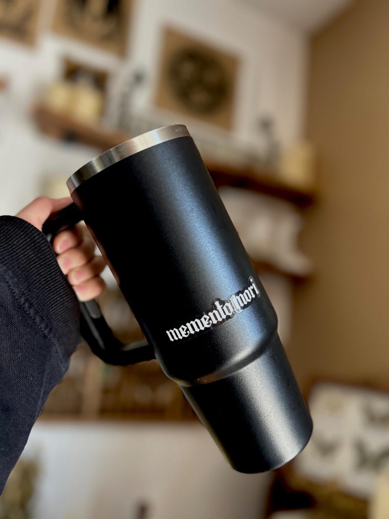 Peut inclure: Mug de voyage en acier inoxydable noir avec une poign&eacute;e noire et une inscription blanche qui dit "memento mori".