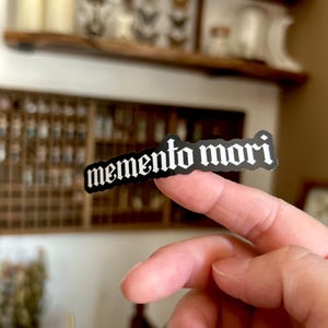 Peut inclure: Un autocollant noir et blanc avec le texte "memento mori" dans une police gothique.