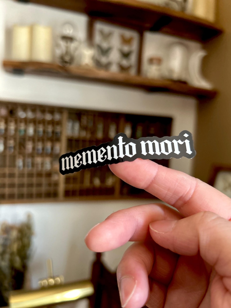 Peut inclure: Un autocollant noir et blanc avec le texte "memento mori" dans une police gothique.