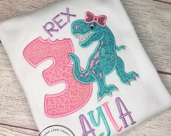 May include: A pink and white number 3 with a turquoise dinosaur wearing a pink bow. The dinosaur is biting the number 3. The name Ayla is embroidered in purple, turquoise, and pink below the dinosaur. The text "Cuba Lake Crafts CLC Embroidery & Design Studio" is in a circle below the name.