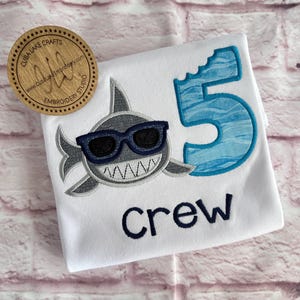 May include: White t-shirt with a blue and gray embroidered shark wearing sunglasses, a blue number 5 with a bite taken out of it, and the word "crew" embroidered in blue.